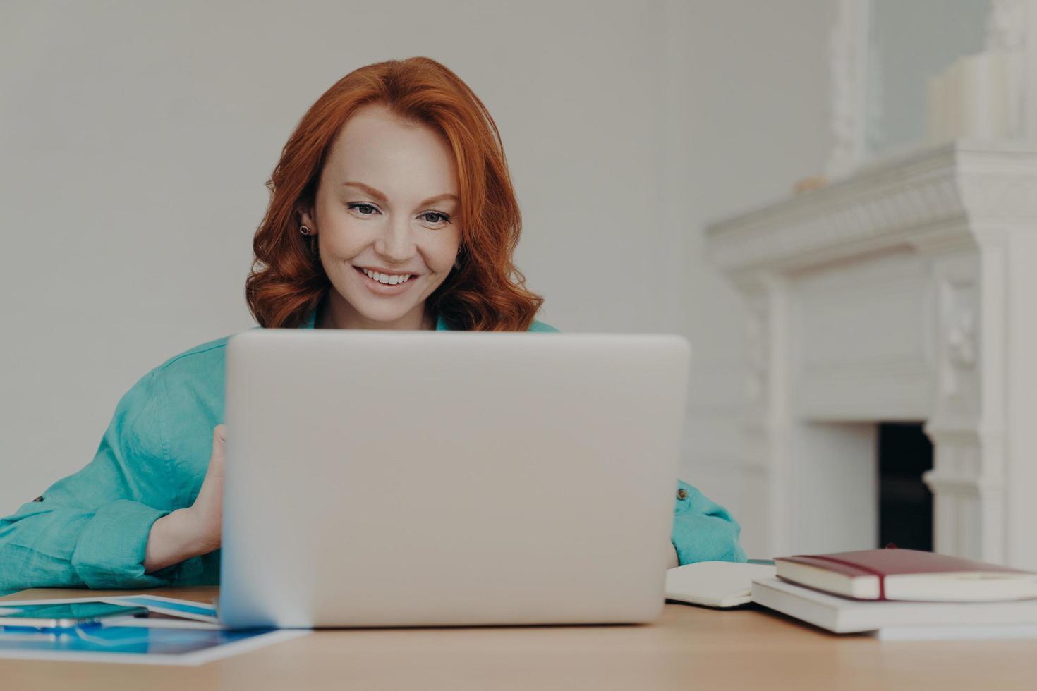 Focused cheerful redhead successful businesswoman does research, makes business project, sits in front of laptop computer at home office desk, uses software application, has happy smile on face photo