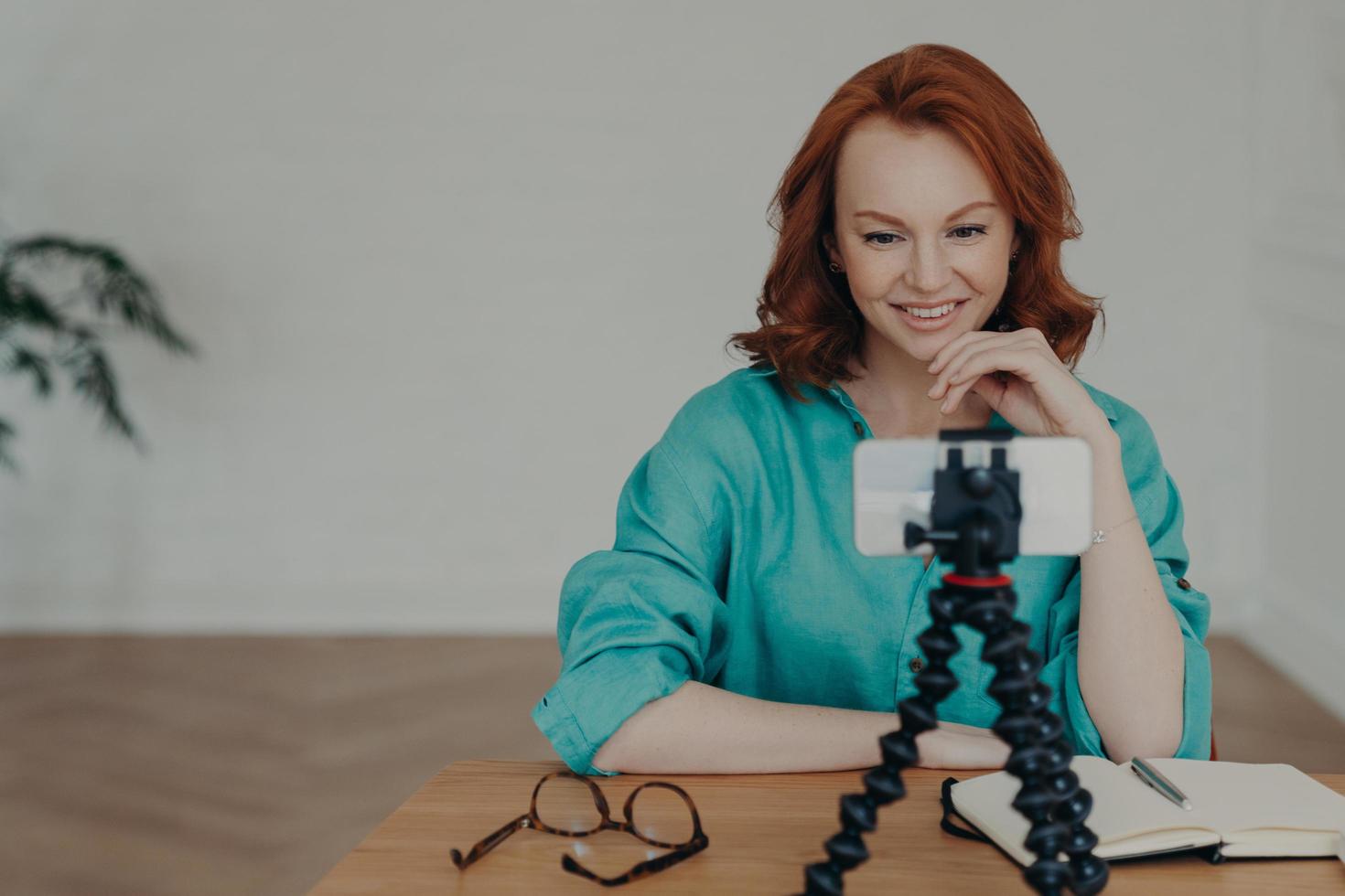 Happy young ginger woman shoots video in front of smartphone videocamera on tripod, gives recommendations how to start own business, has personal vlog, speaks on video call about online training photo