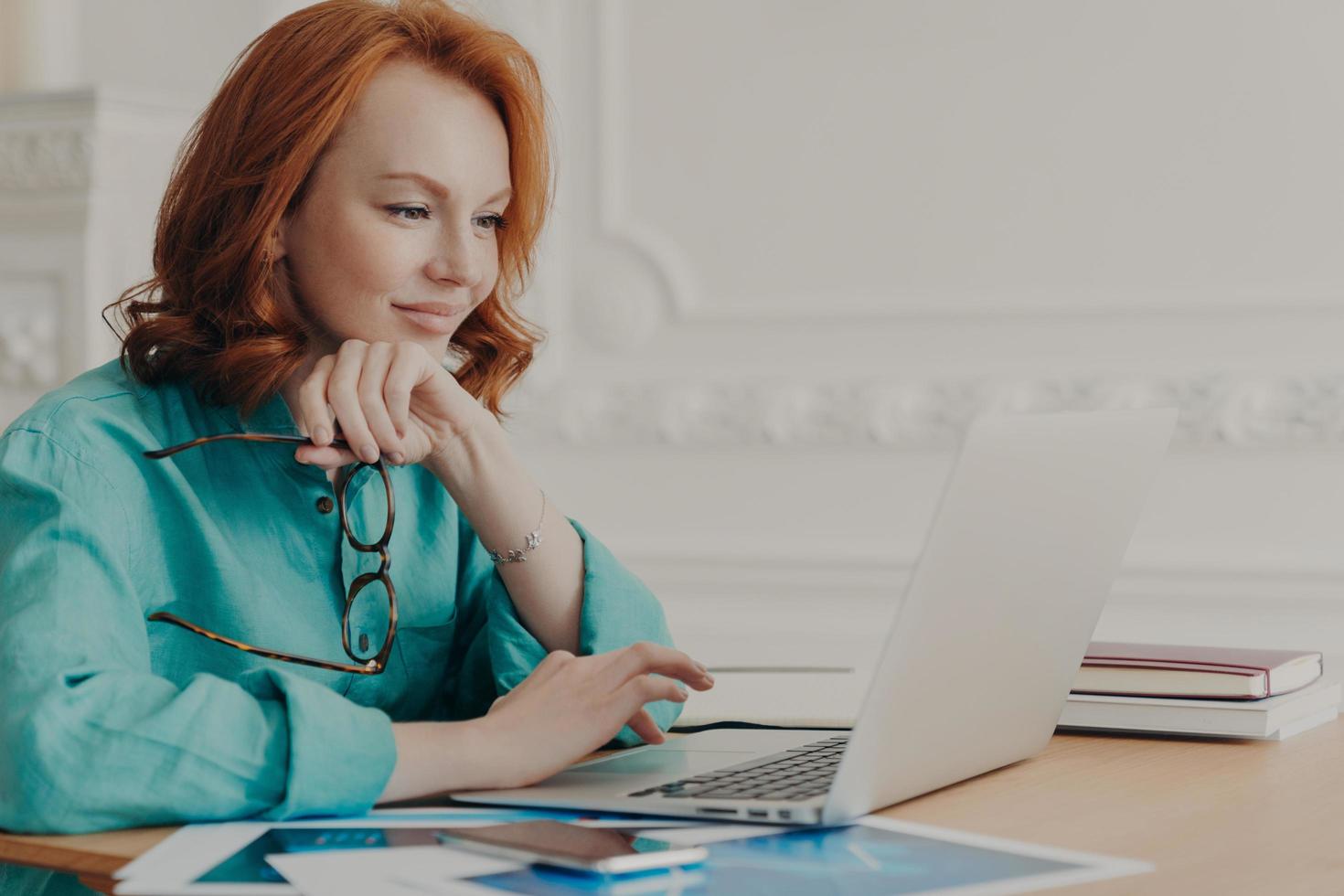 Busy redhead woman entrepreneur works on modern laptop, holds spectacles, poses in coworking space, reads email, received good business news, does remote job from home, enjoys informal chat. photo