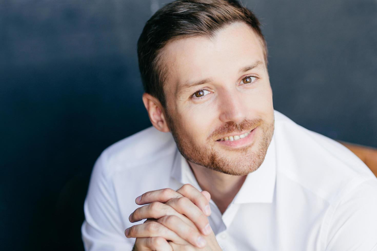 Close up portrait of handsome unshaven male with positive smile, keeps hands together, feels successful as works in business sphere, wears white shirt, isolated over dark background. Emotions concept photo