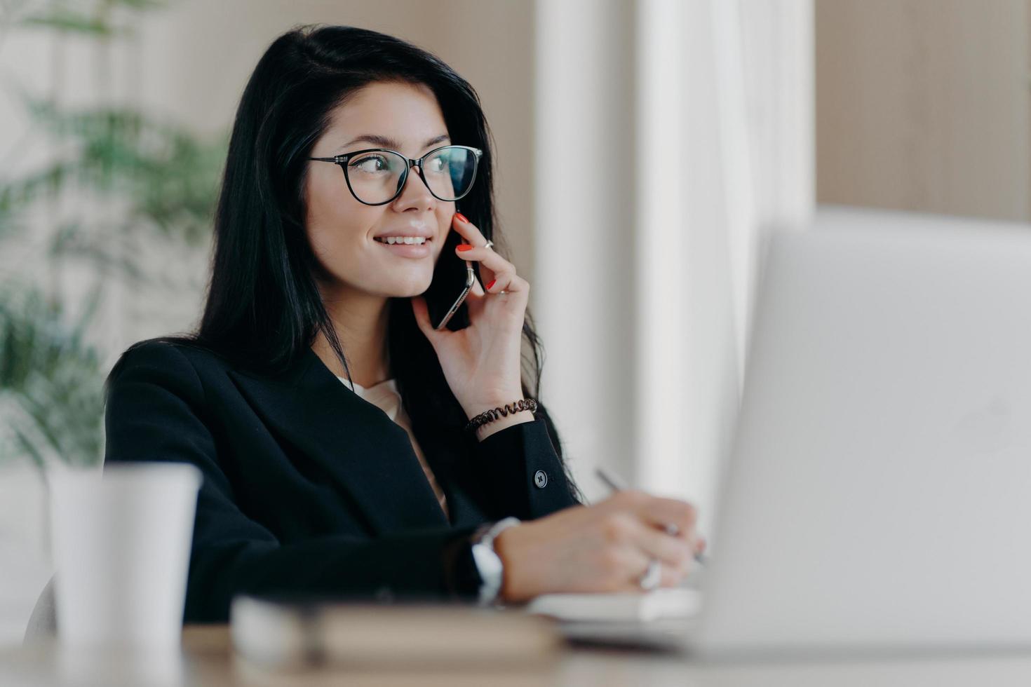 Elegant female entrepreneur with satisfied expression has telephone conversation, checks agenda while working on modern laptop computer, writes down notes in notepad, poses in coworking space photo