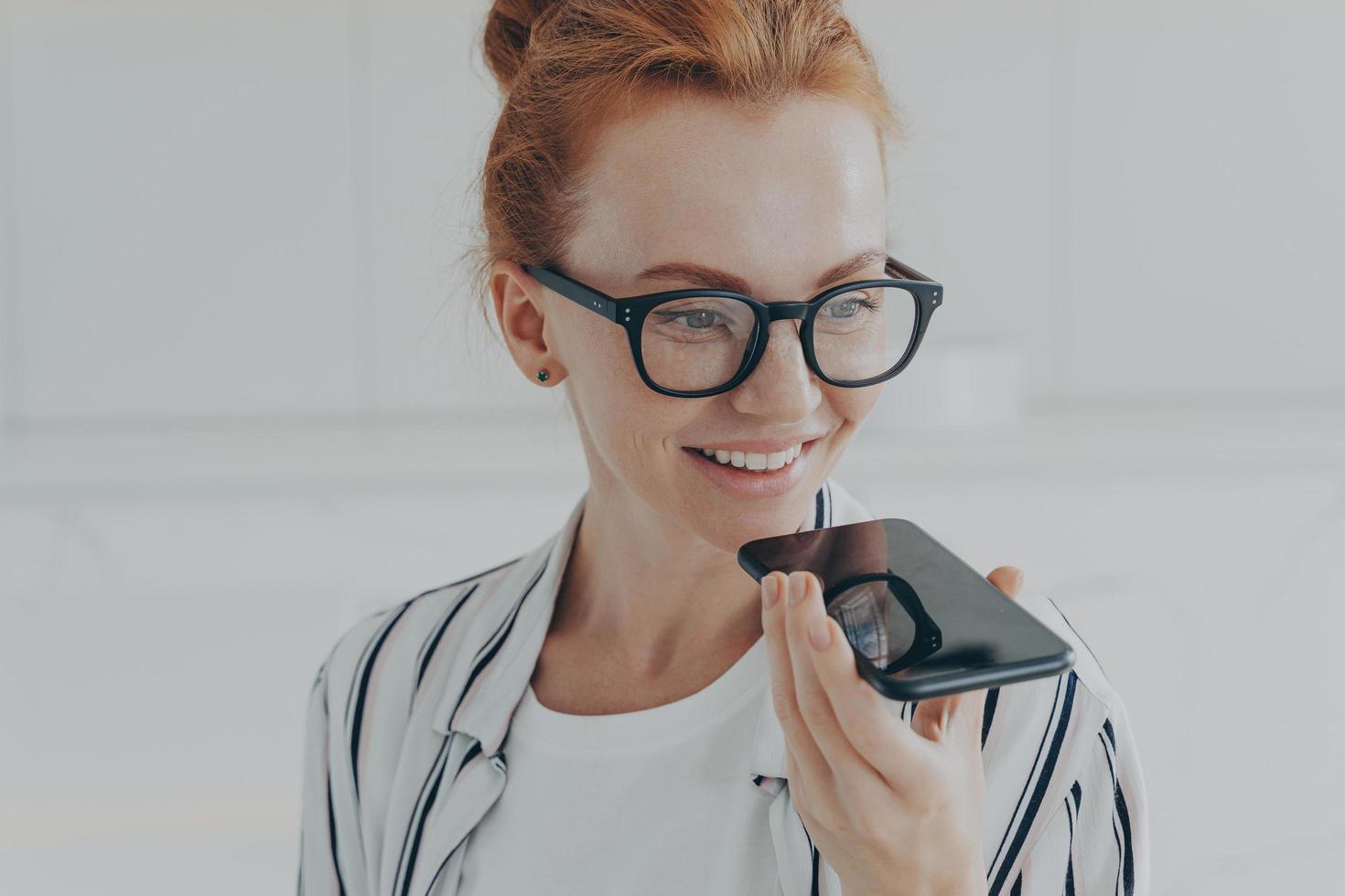 mujer pelirroja con gafas transparentes habla con un asistente virtual que sostiene el teléfono móvil cerca de la boca foto