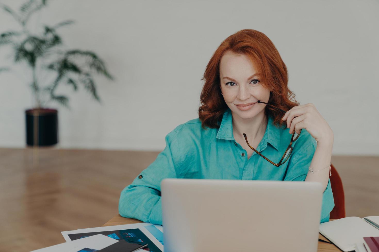 Beautiful happy busy female freelance worker with ginger hair, works online, uses laptop computer, prepares financial report at home, uses videoconferencing app, talks distantly with colleagues photo