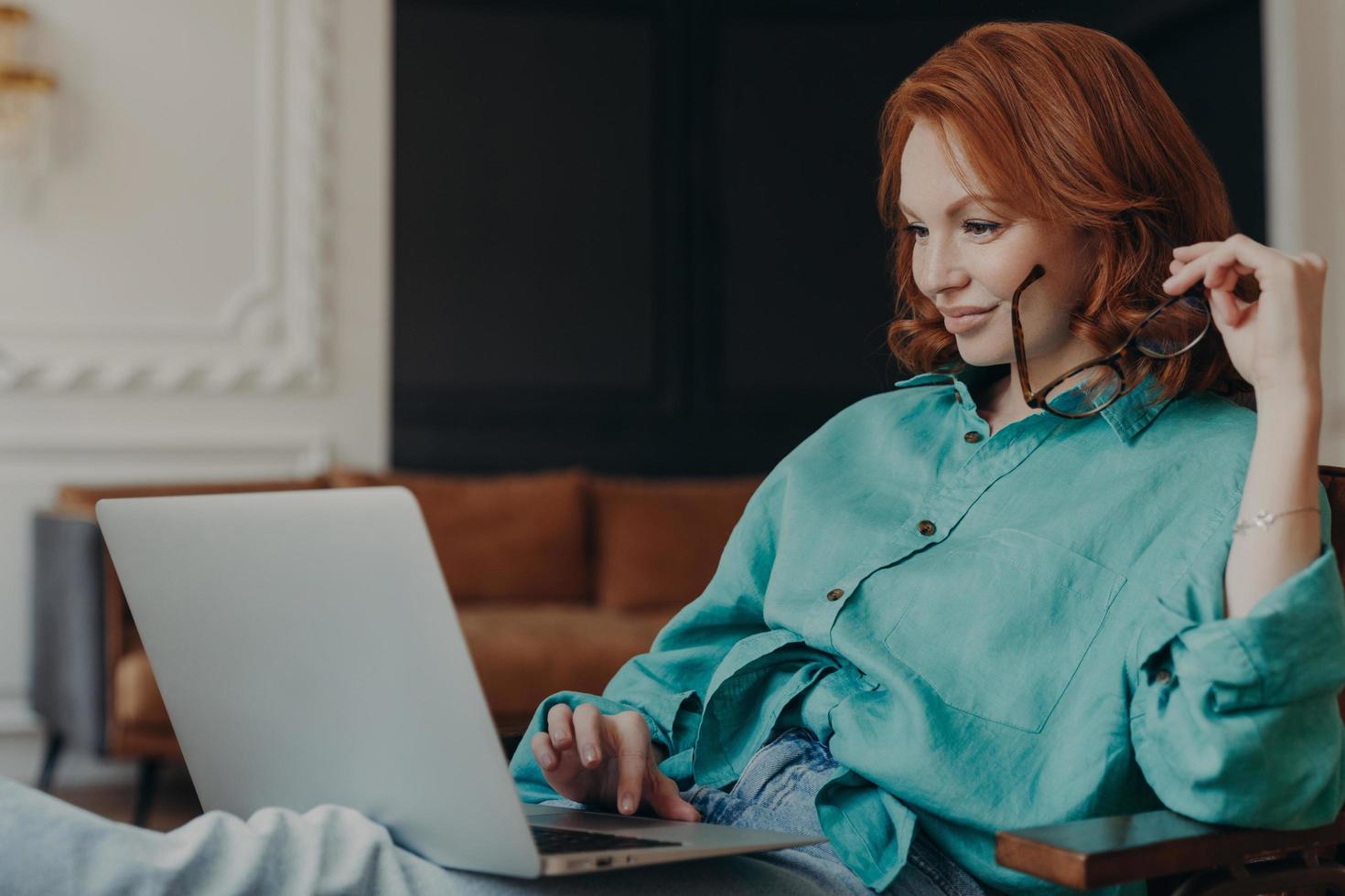 Pleased young redhead woman sits on comfortable armchair at home, does remote work, reads news in social networks, busy with computer job, concentrated in monitor of laptop. Modern technologies photo