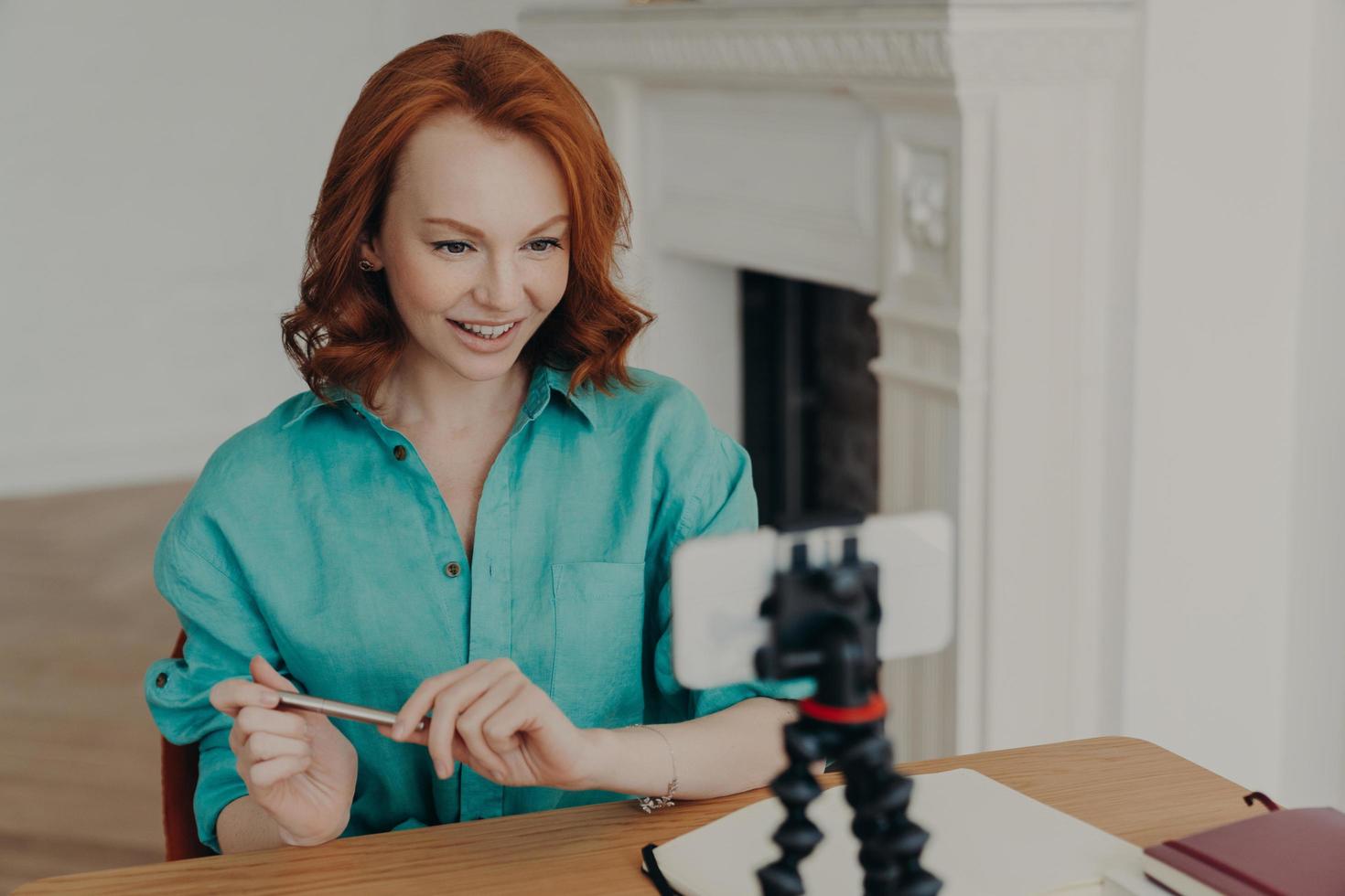 Social media influencer blogger keeps mobile phone on tripod, looks gladfully at camera, streams online podcast, shoots vlog, sits at desktop, has online communication or chat, uses special app photo