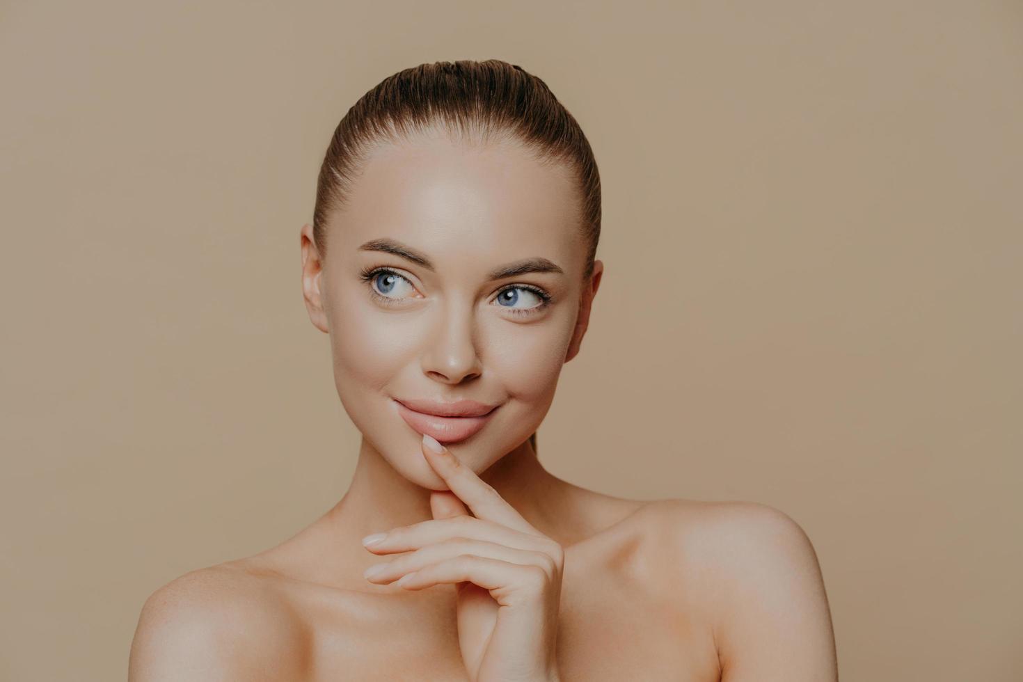 Pensive attractive young European woman with white healthy skin, looks aside, has naked body, undergoes beauty treatments, poses against beige background, has combed dark hair, bare shoulders photo