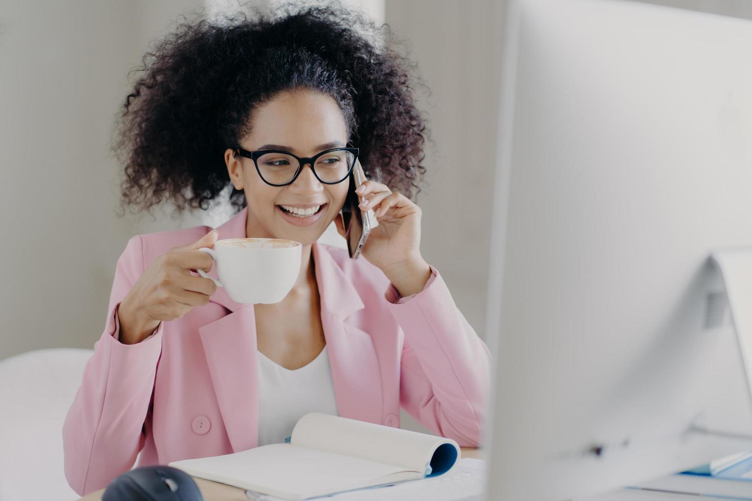 Photo of attractive smiling female entrepreneur enjoys aromatic beverage, holds white mug of drink, has telephone conversation, dressed in formal outfit, poses at workplace, wears transparent glasses