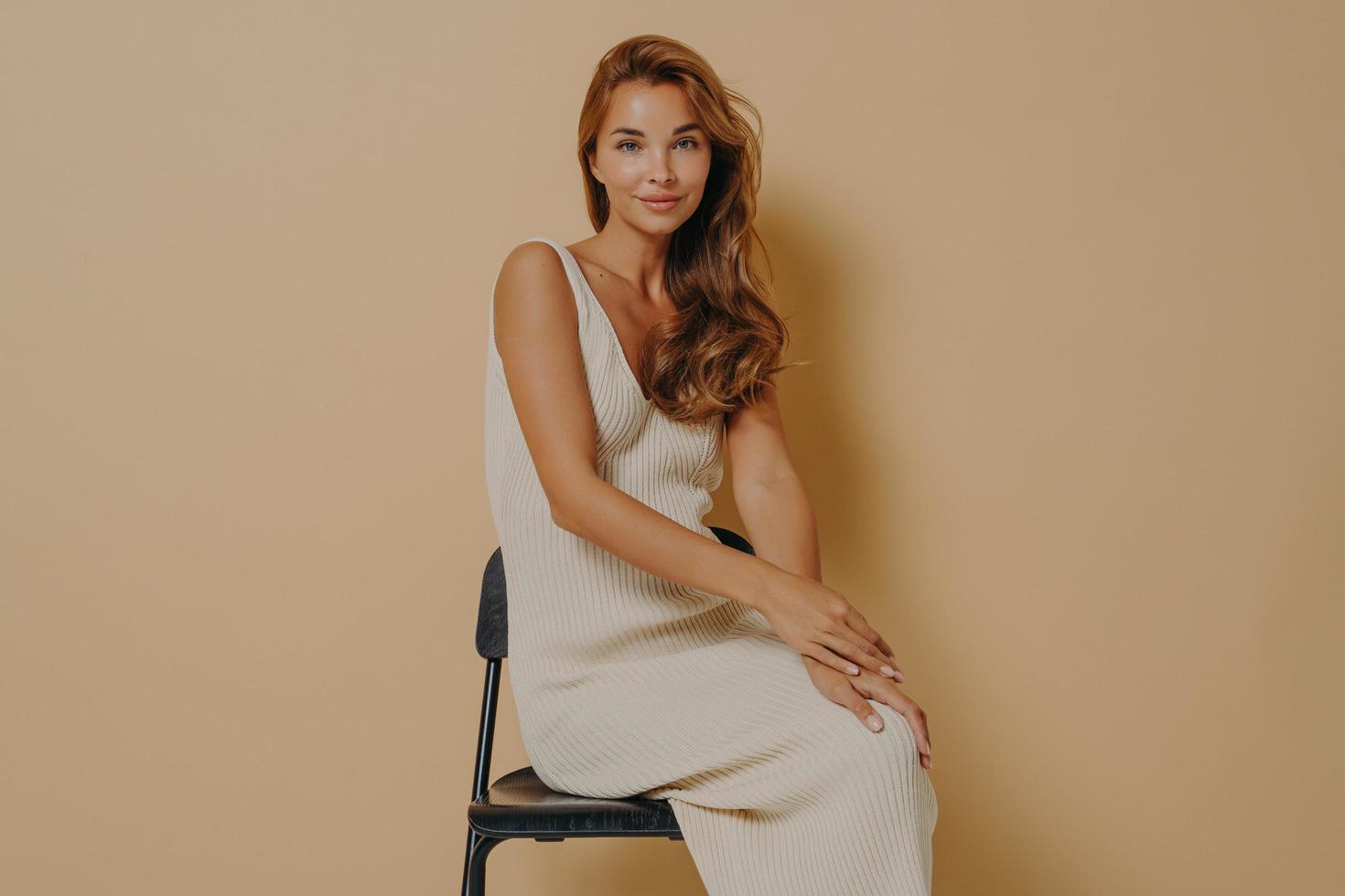 Sideways shot of beautiful lady with wavy hair poses on chair against beige background photo