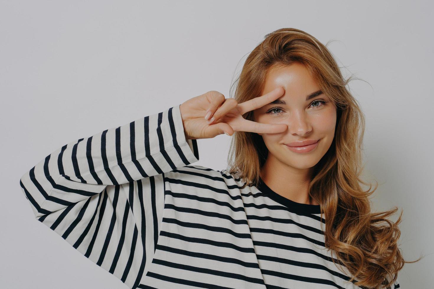 Happy young European woman makes peace disco gesture over eye v sign wears casual striped jumper photo
