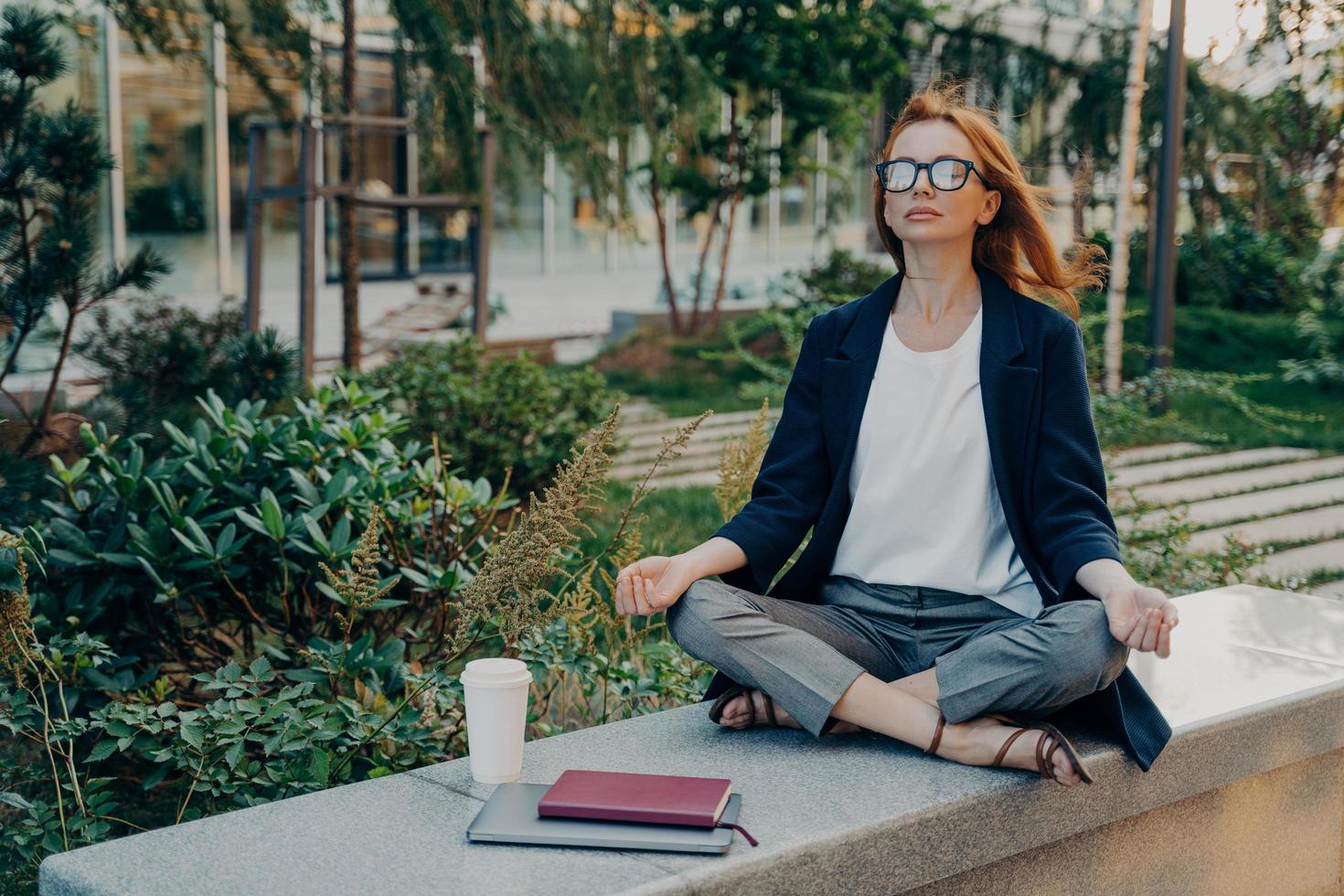 Peaceful business woman holding hands in mudra gesture, sitting in lotus pose and meditating in park photo