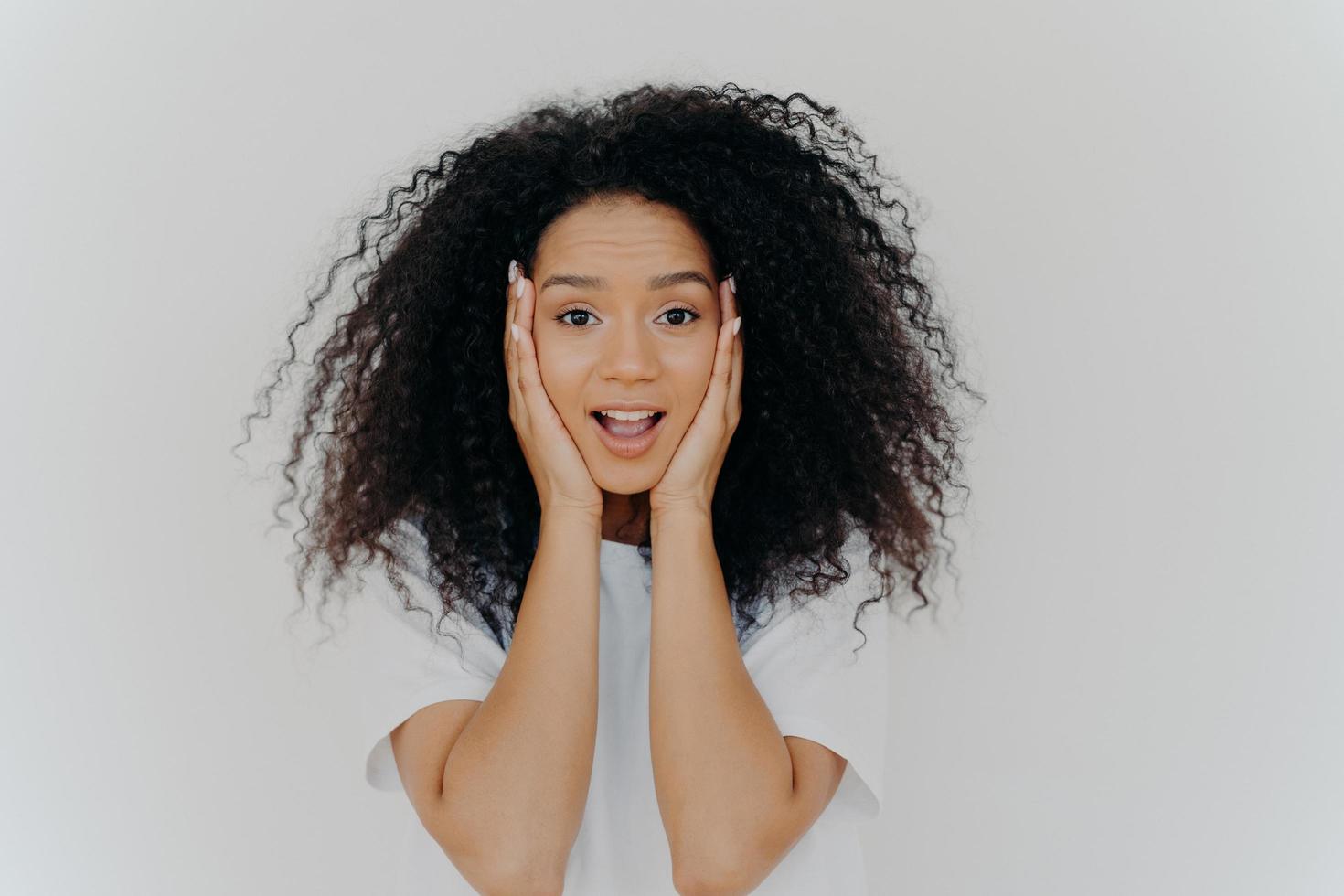 foto de mujer alegre sorprendida con corte de pelo afro, mantiene ambas manos en las mejillas, tiene belleza natural, abre la boca, no puede creer en noticias emocionantes, usa camiseta, posa contra fondo blanco.