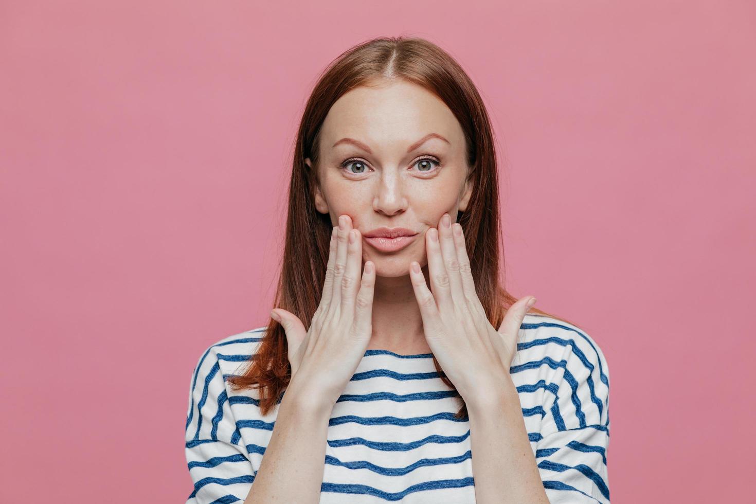 la foto de una mujer bonita pecosa mantiene las manos cerca de los labios, tiene un maquillaje mínimo, piel sana, cabello castaño, demuestra belleza natural, vestida con un traje a rayas, aislada sobre un fondo rosa.