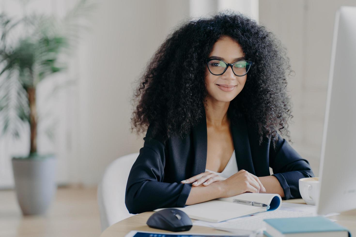 Positive female director with Afro curly hair, rewrites information in notepad from computer, wears optical glasses and formal wear, drinks coffee, poses at desktop, prepares working schedule photo
