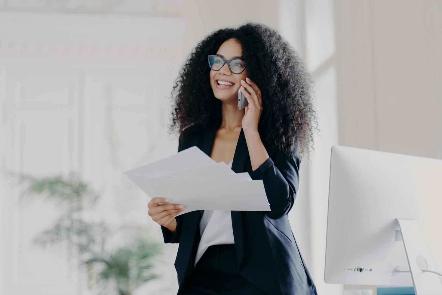 Pleased successful businesswoman with Afro hairstyle wears spectacles to provide eyes protection, holds documents, makes telephone call to business partner, stands in cabinet near workplace. photo