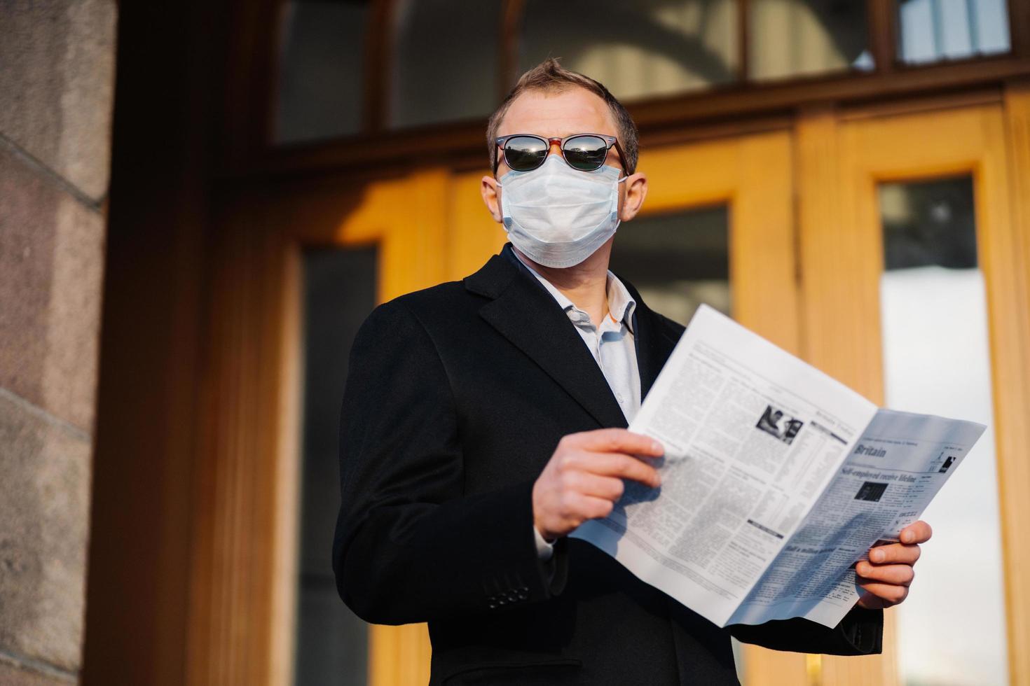 hombre caucásico con gafas de sol, máscara médica y traje negro formal, se para cerca del edificio al aire libre, sostiene el periódico en las manos, posa en la calle, camina en la ciudad en cuarentena, descubre noticias de la prensa foto