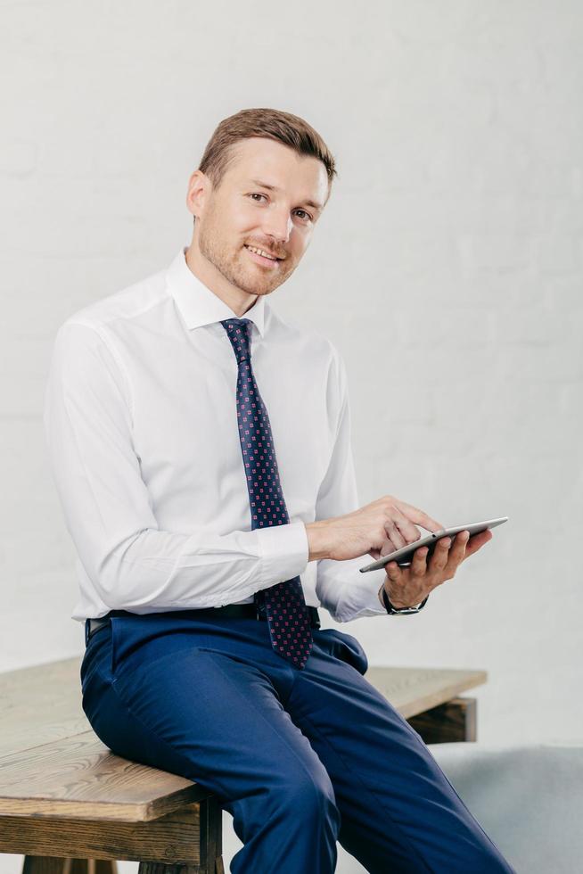 Smiling young businessman in elegant clothes happy to recieve income message on modern tablet computer, sits at wooden office desk in his cabinet, poses against white wall. Business concept. photo