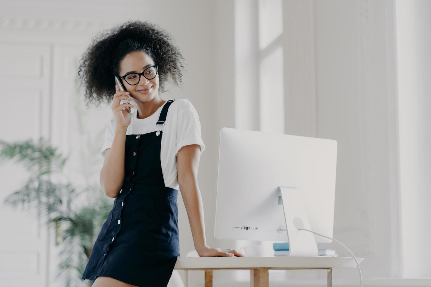 Pleasant looking African American female has telephone conversation, asks for advice before job interview, dressed in stylish clothes, poses near workplace with modern technology. Communication photo