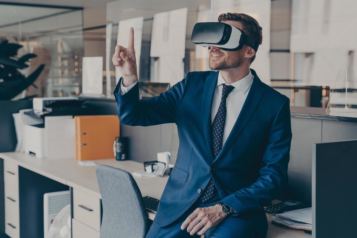 Enjoyed young businessman pointing with forefinger up in air with virtual reality glasses on head photo