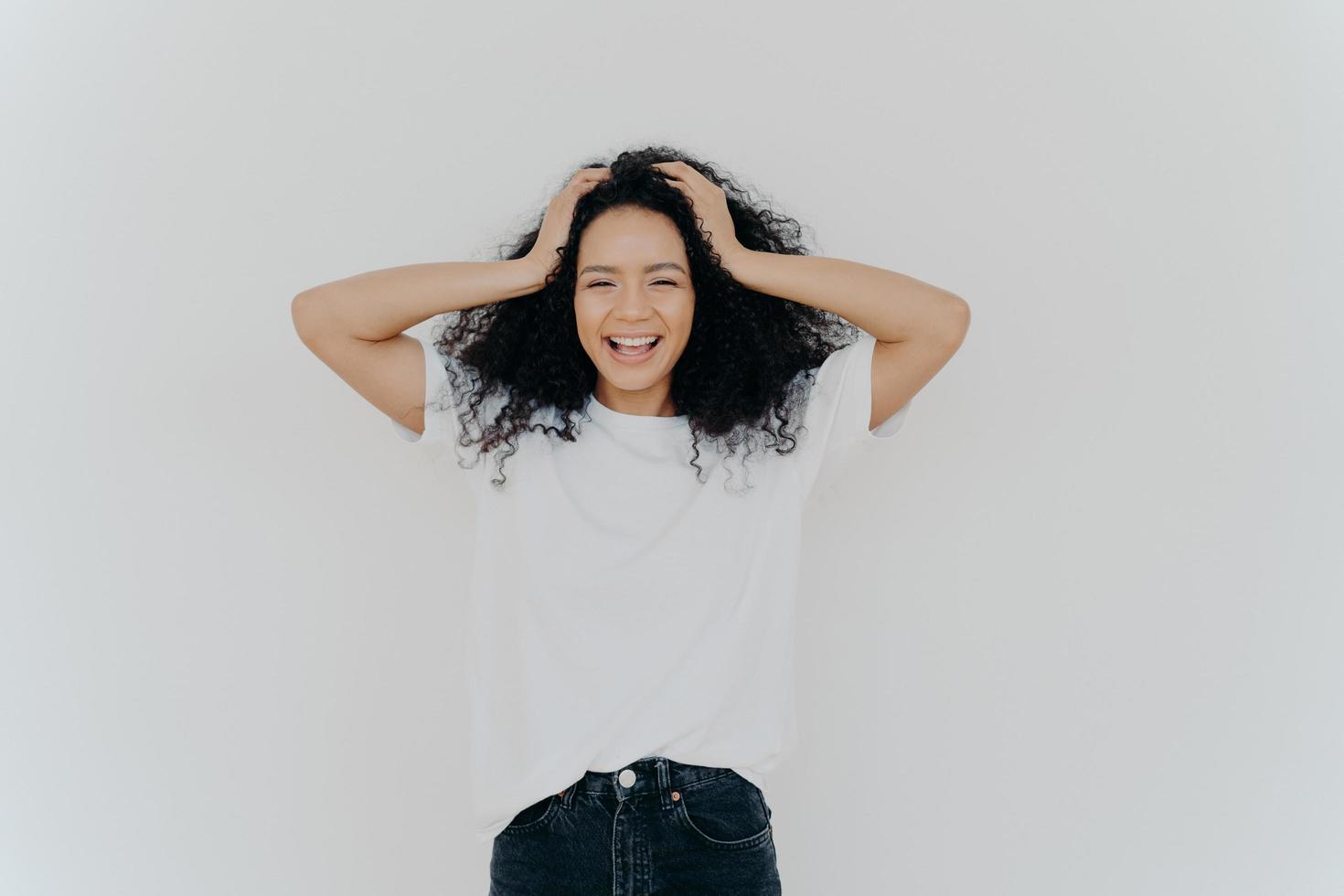 Photo of overjoyed curly haired woman laughs happily, has fun, dressed in white t shirt and jeans, smiles broadly, isolated over white background. People, emotions, happiness, ethnicity concept