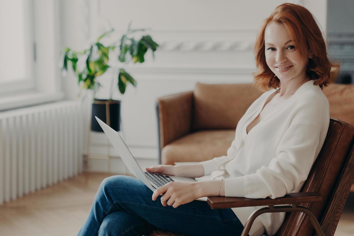Sideways shot of happy redhead young woman sits in comfortable armchair with laptop computer, works remotely, checks email and reads notification, browses useful website and installs application photo