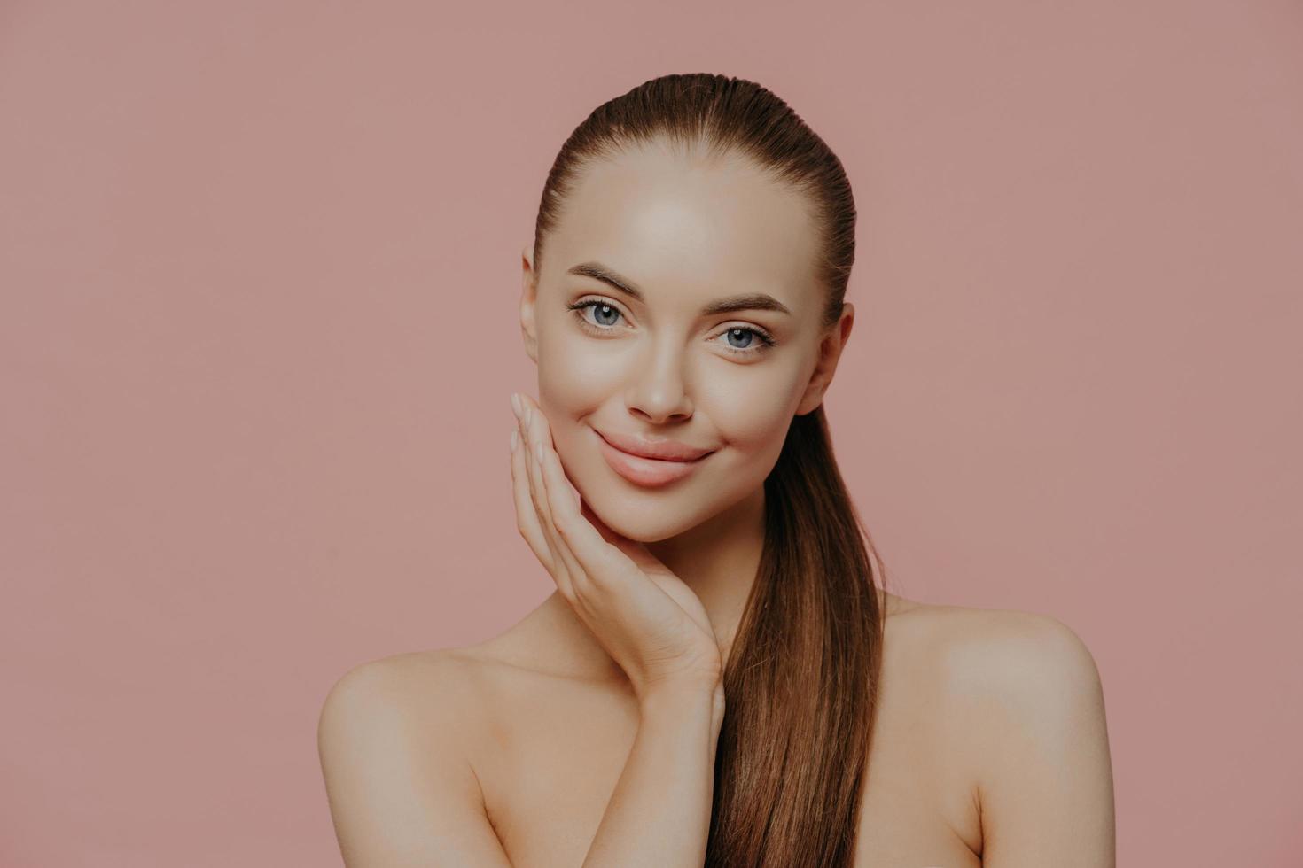 Portrait of good looking female has long straight hair combed in pony tail, stands shirtless, has clean perfect skin, satisfied with her natural beauty, poses indoor against pink background. photo