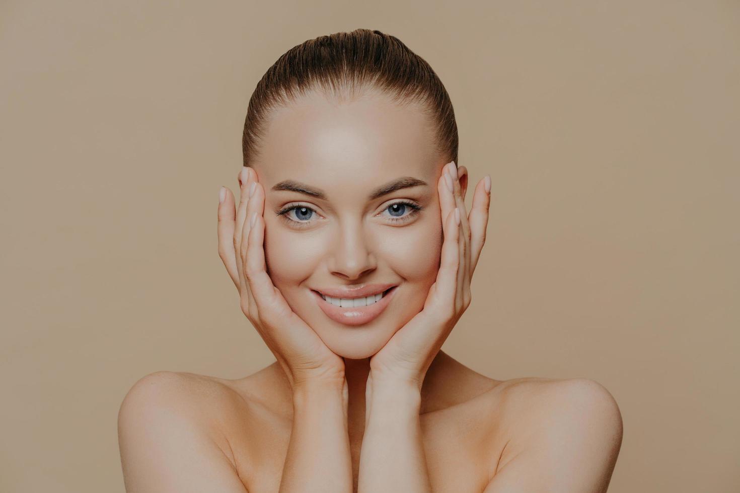 concepto de belleza y cuidado de la piel. una foto en la cabeza de una mujer hermosa y alegre toca la cara suavemente, tiene una sonrisa perfecta, una piel sana después de limpiar o aplicar una máscara facial, aislada sobre un fondo beige