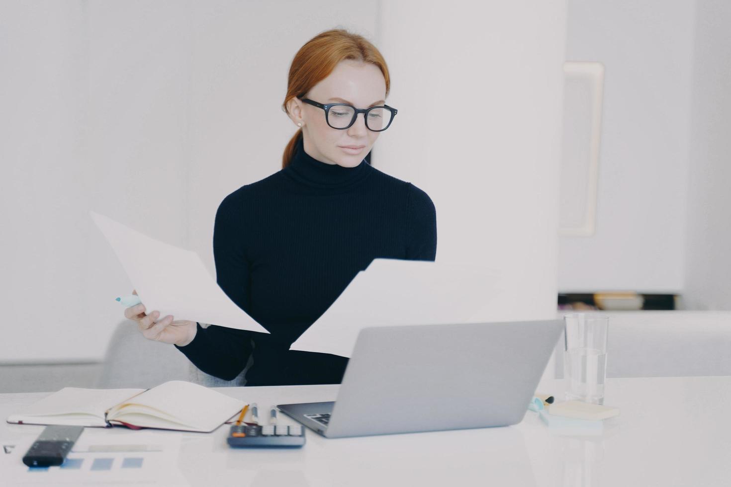 Concentrated woman secretary is working at laptop and holding document in office. photo