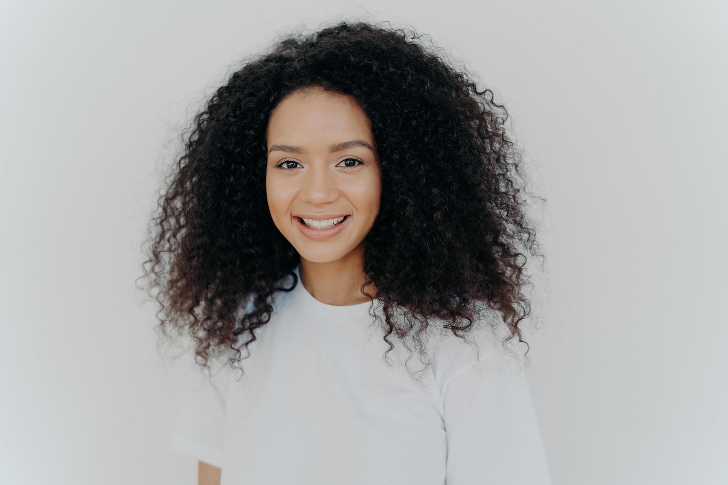 Happiness, wellbeing and positive emotions concept. Headshot of pleasant looking lady with crisp hair, smiles and laughs, has healthy skin, wears casual t shirt, models against white studio wall. photo