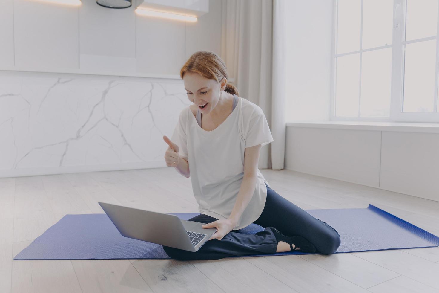 Positive woman sitting on floor having yoga lesson in front of laptop. Girl shows thumbs to coach. photo