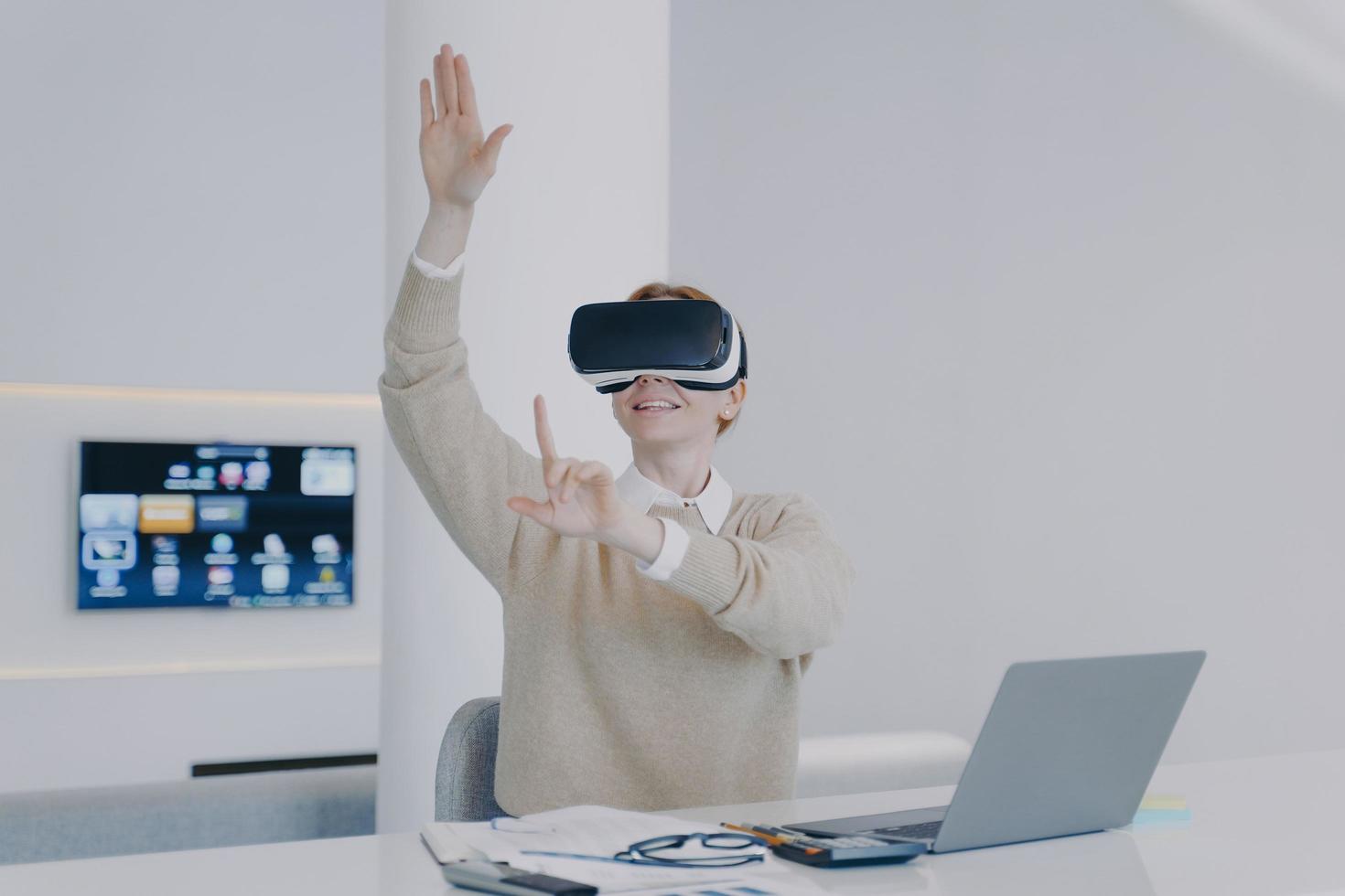 Young white woman in VR goggles working on project in cyberspace in front of laptop in office. photo