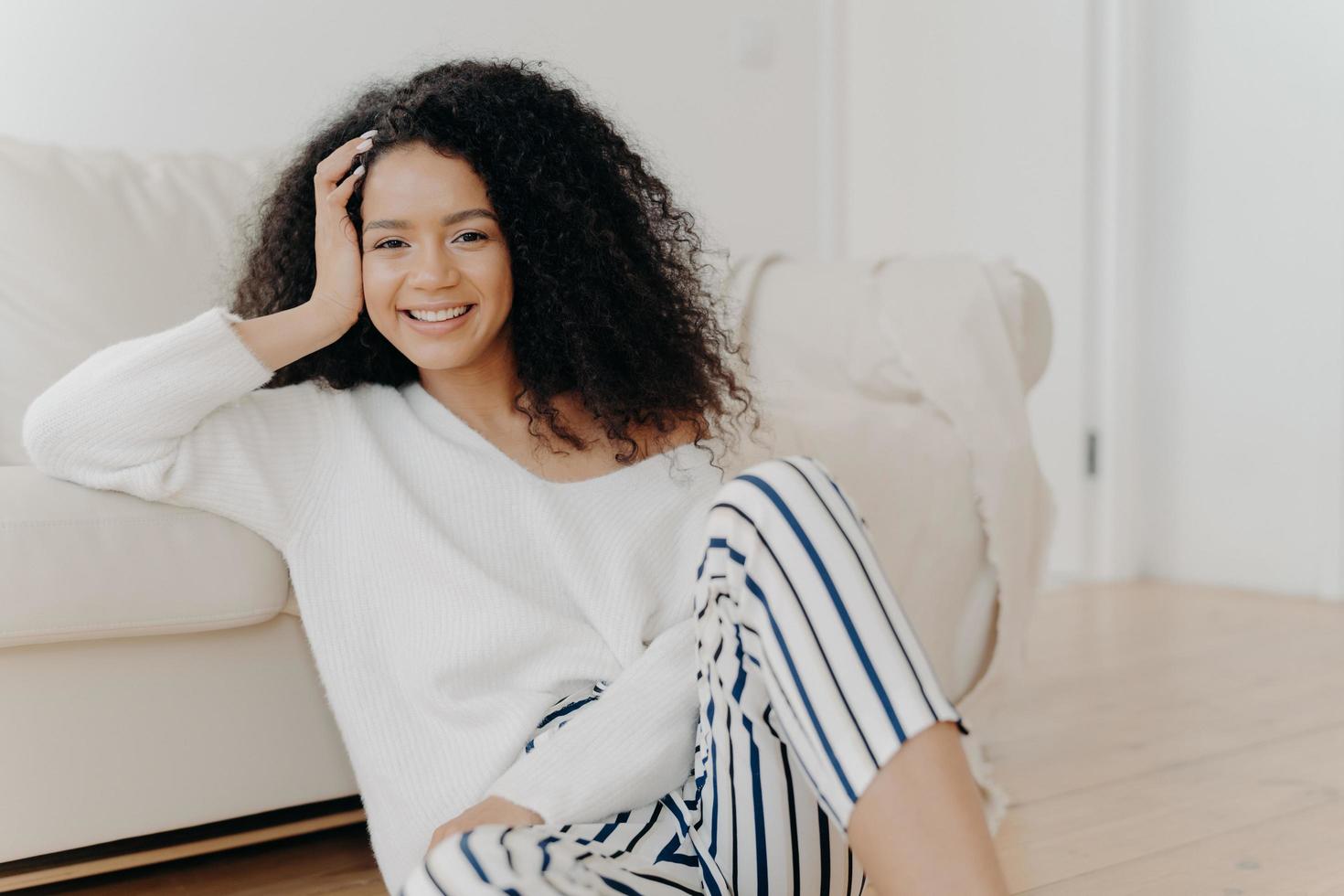 Lovely dark skinned woman with crisp hair, sits on floor near sofa, wears white sweater and striped trousers, enjoys positive emotions, enjoys weekend at home. People, rest, happiness concept photo