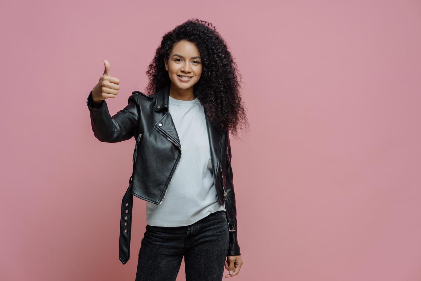 Pleased curly haired woman keeps thumb up, makes approval gesture, dressed in white t shirt, stylish leather jacket and jeans, poses against pink background. Body language concept photo