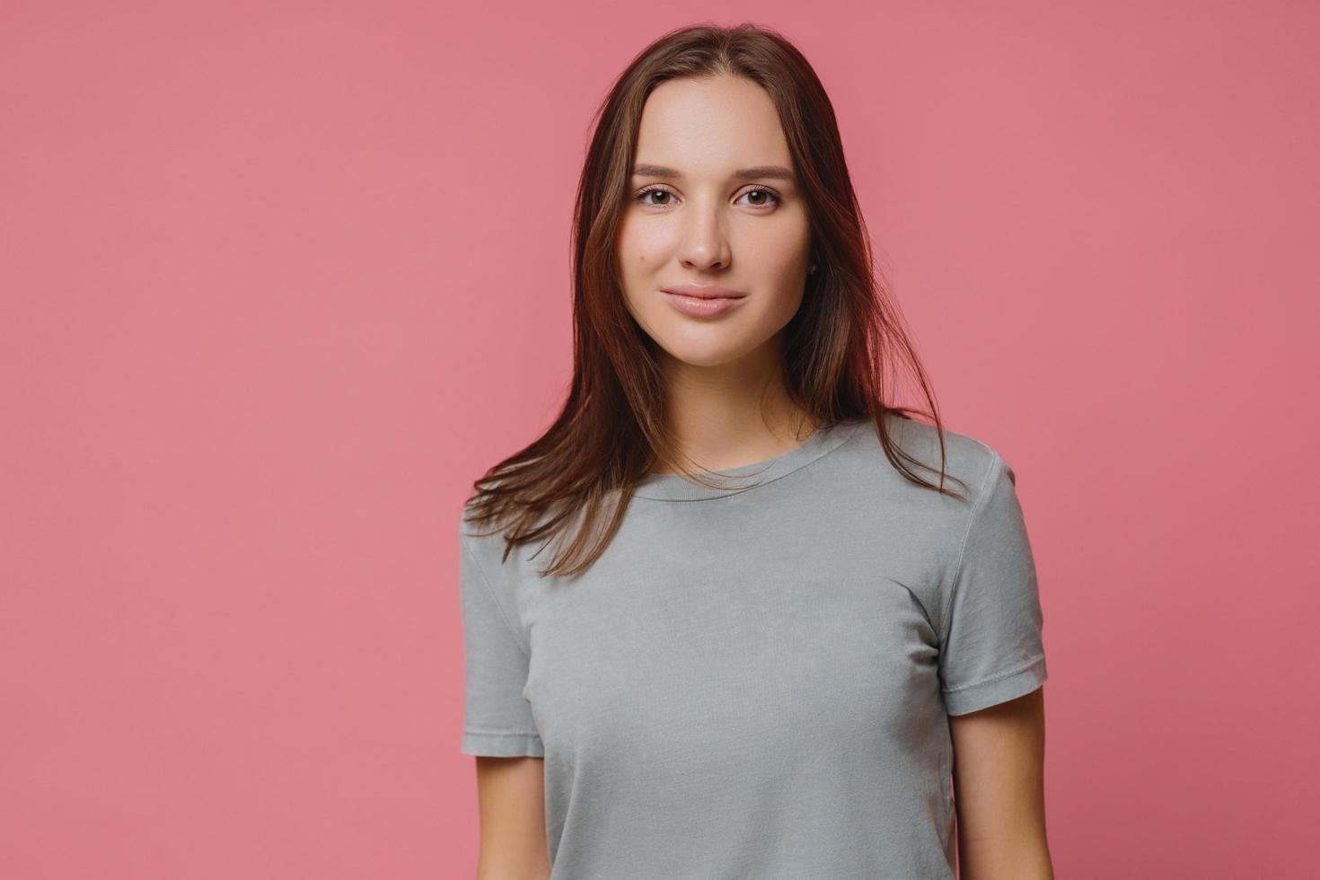 Adorable millennial girl with straight dark hair, wears casual t shirt, looks seriously at camera, has healthy skin, poses over pink background. Pleasant looking woman has talk with someone. photo