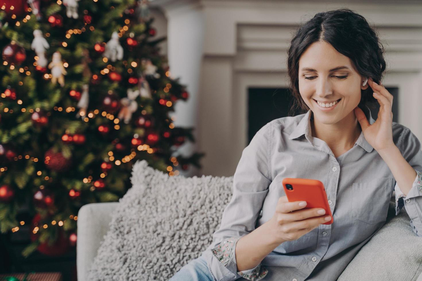 relajada mujer italiana feliz sentada en un acogedor sofá en casa con teléfono durante las vacaciones de Navidad foto