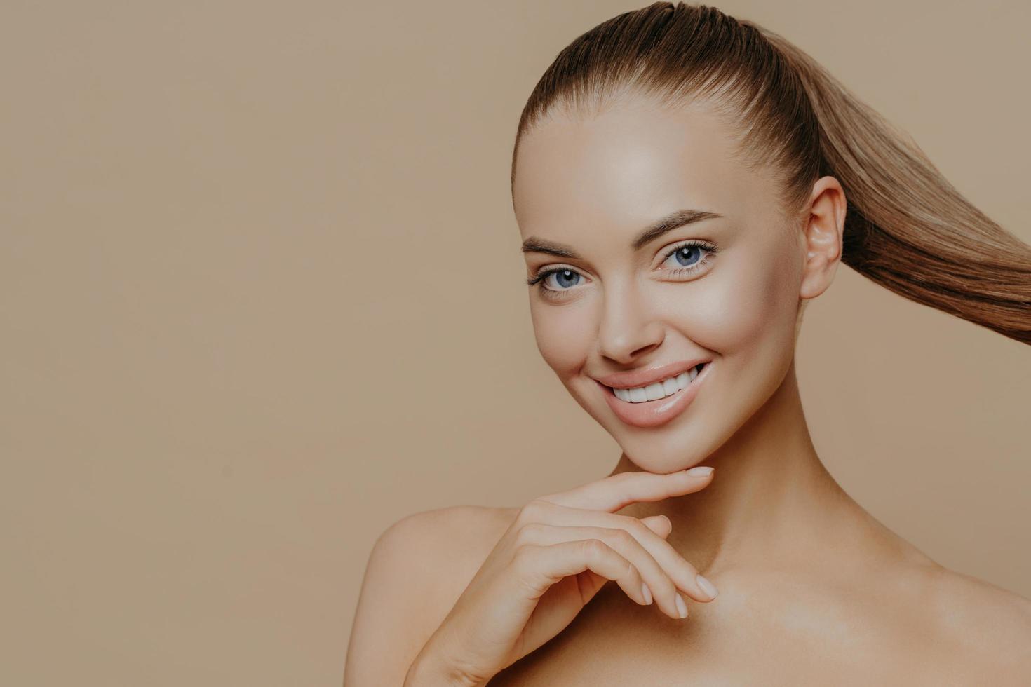 Tranquil undressed woman with healthy perfect skin, dark hair combed in pony tail, enjoys facial treatments, has well cared complexion, stands against beige studio wall, empty space. Skin care concept photo