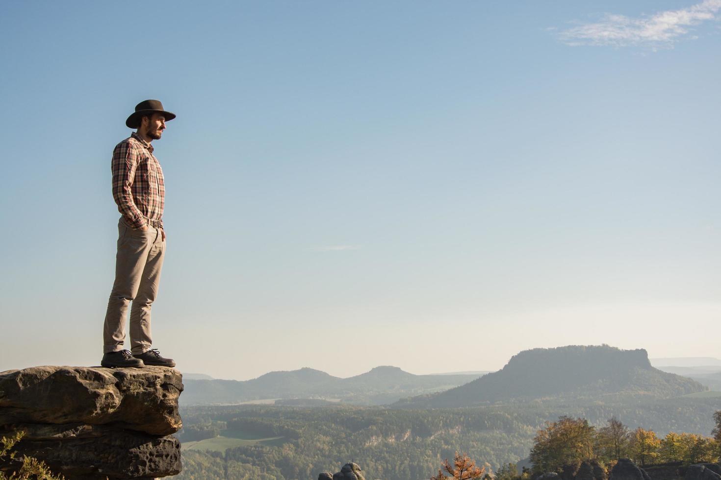 Young bearded male traveler in hat stand on the cliff in sunny day photo