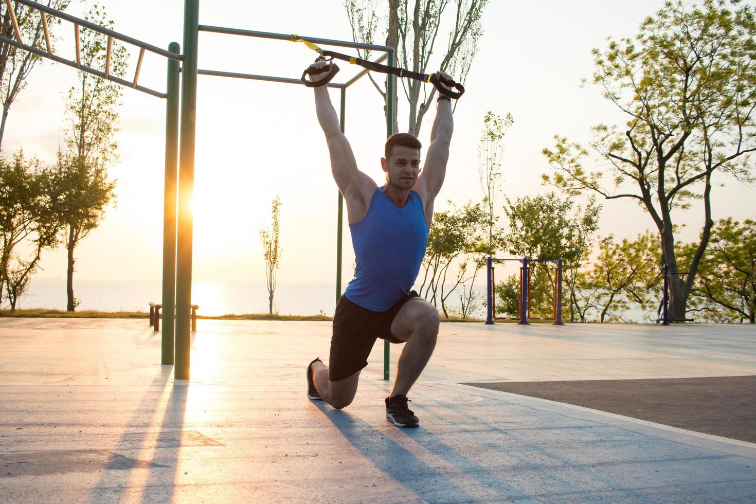 workout with suspension straps In the outdoor gym, strong man training early in morning on the park, sunrise or sunset in the sea background photo