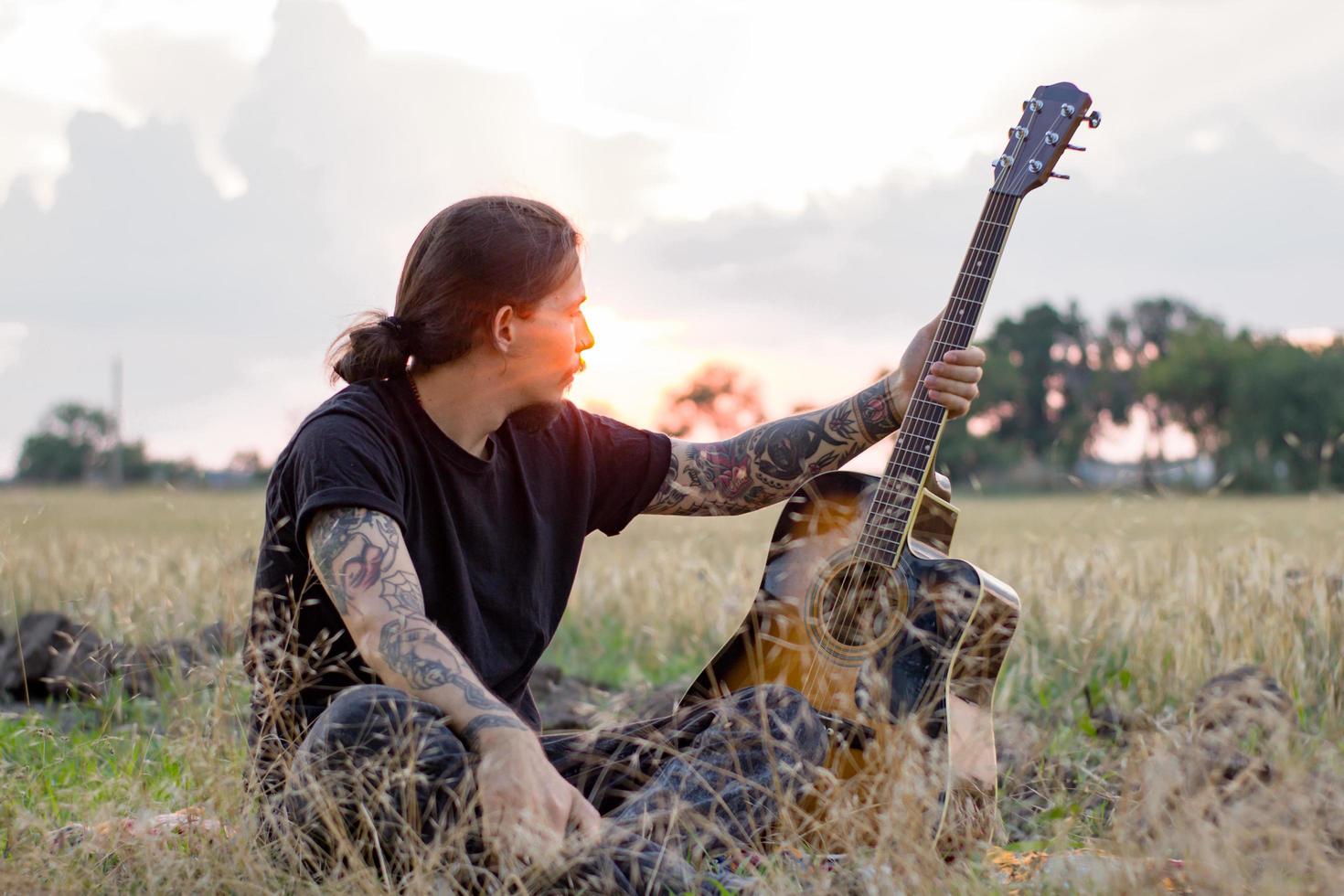 Young tattooed male play on acoustic guitar in summer fields during beautiful sunset photo