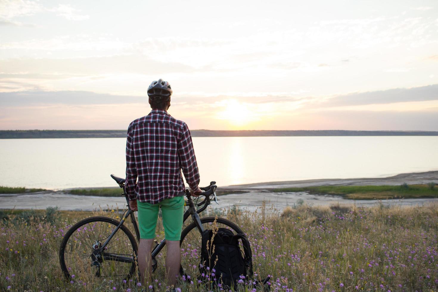 silueta de un hombre con bicicleta de carretera de gira mirando y haciendo una foto de la puesta de sol en el lago en el teléfono celular