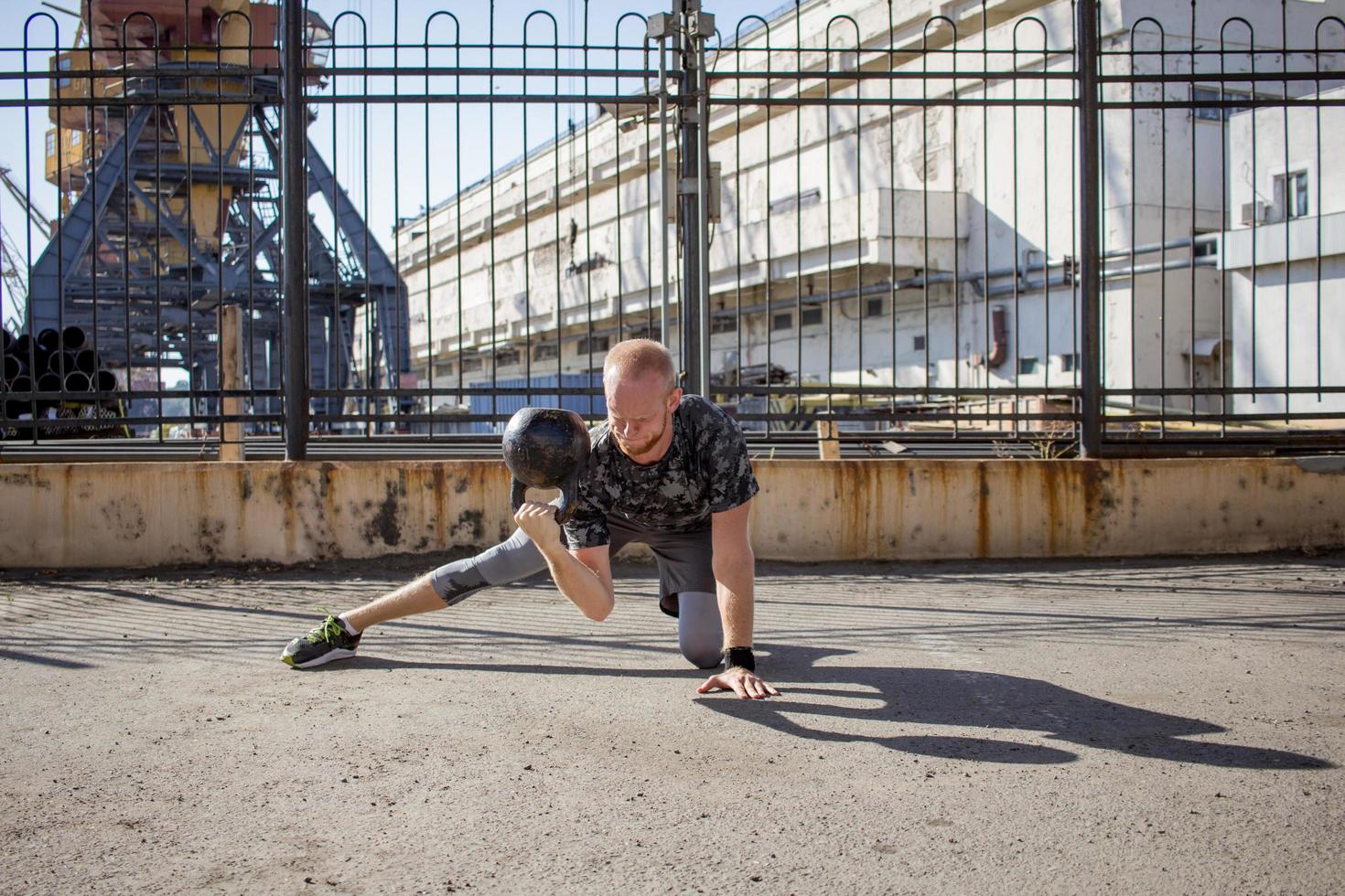 joven atleta masculino barbudo entrenando en zona industrial en un día soleado, ejercicios de campanas al aire libre, fondo urbano foto