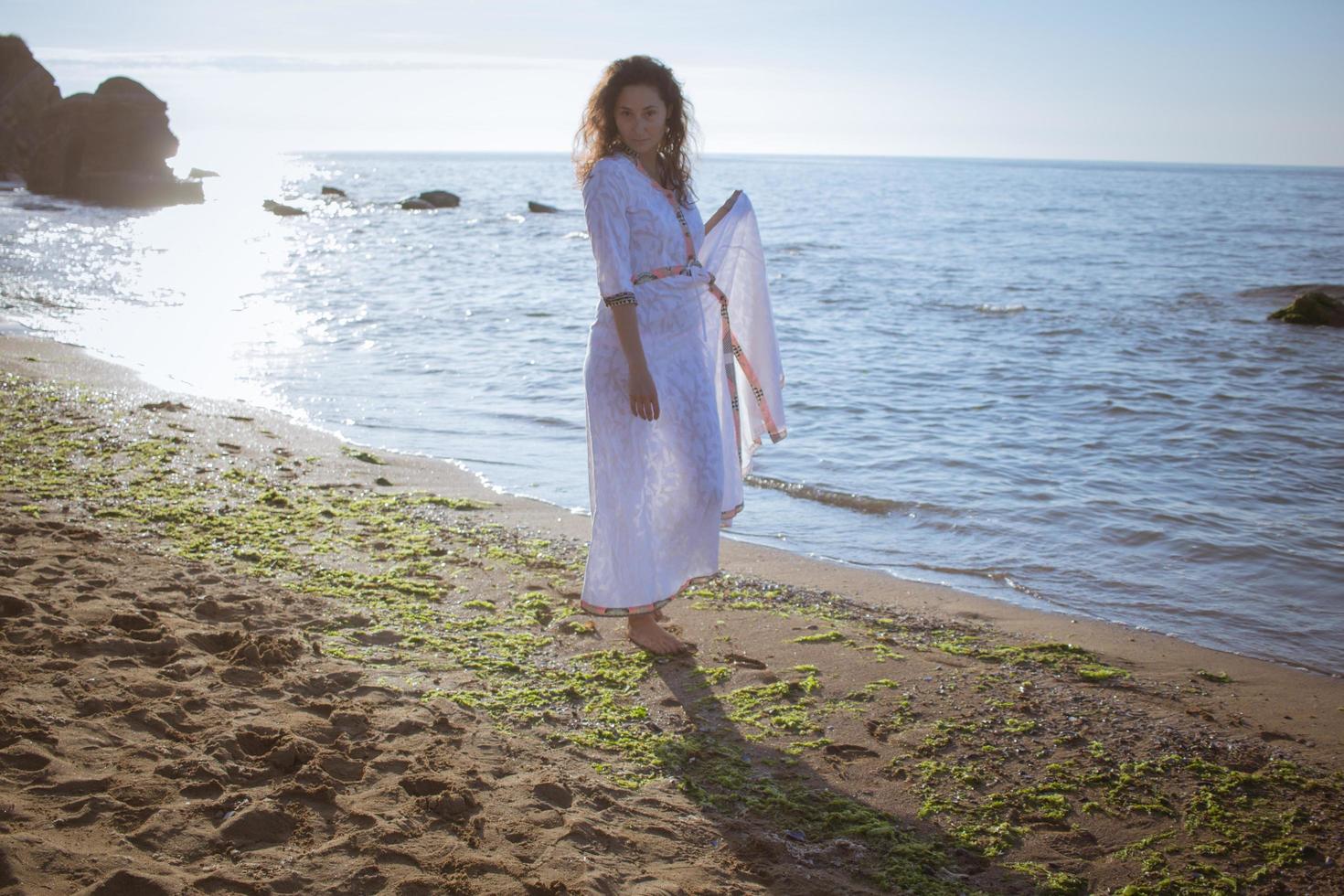 mujer joven caminando en la playa de la mañana en un hermoso vestido blanco. mujer en forma pasando un buen rato durante el amanecer. foto