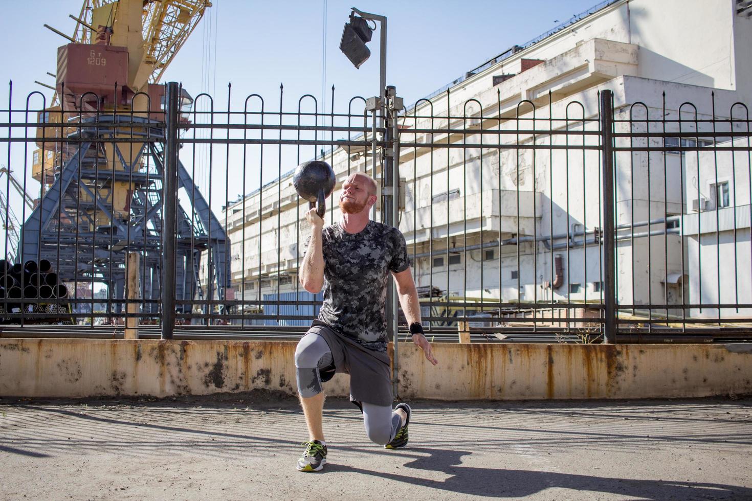 Young bearded male athlete training in industrial zone in sunny day, kettlebells exercises outdoors, urban background photo