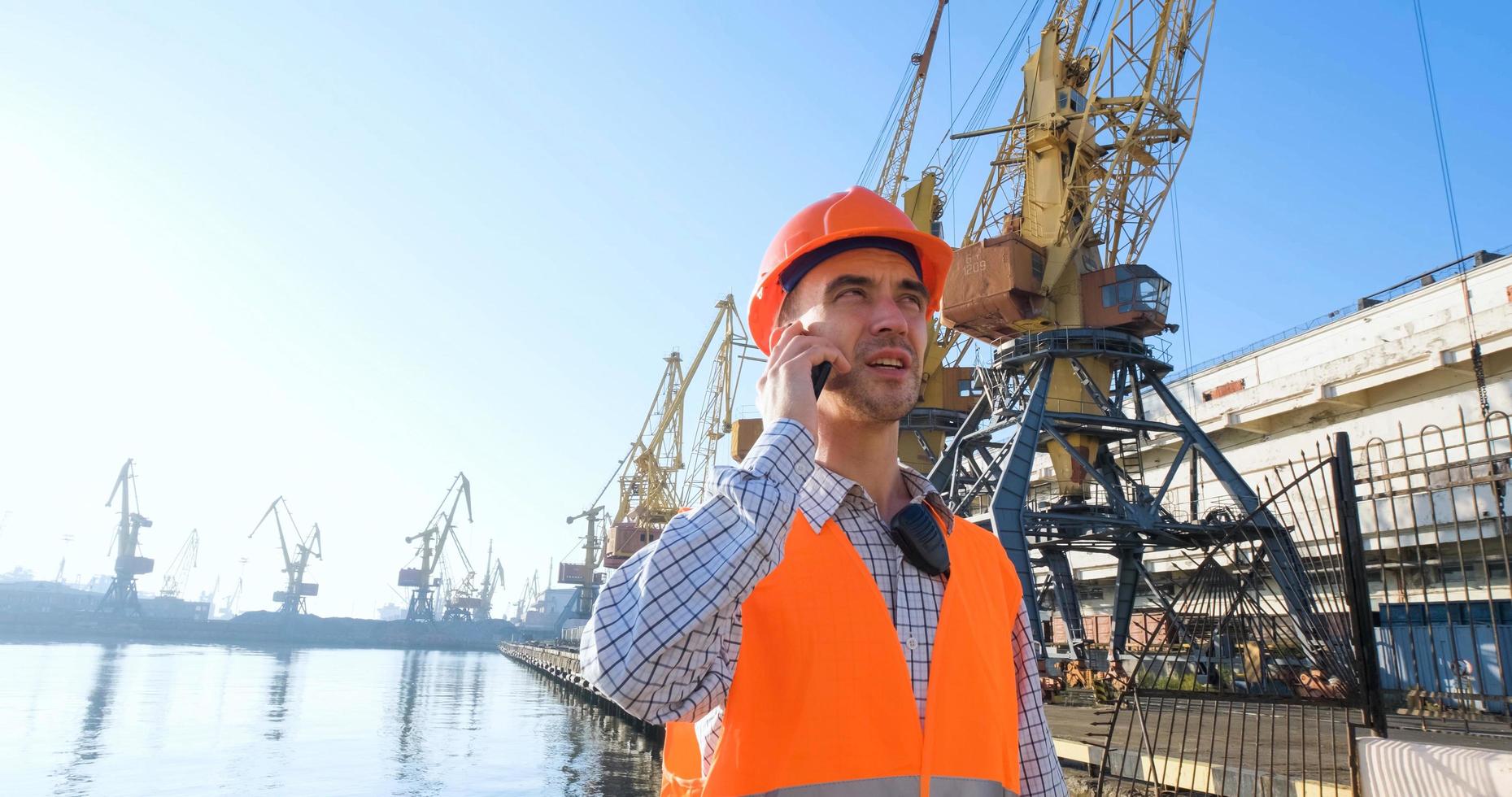 male worker of sea harbor in orange helmet and safety west, cranes and sea background photo
