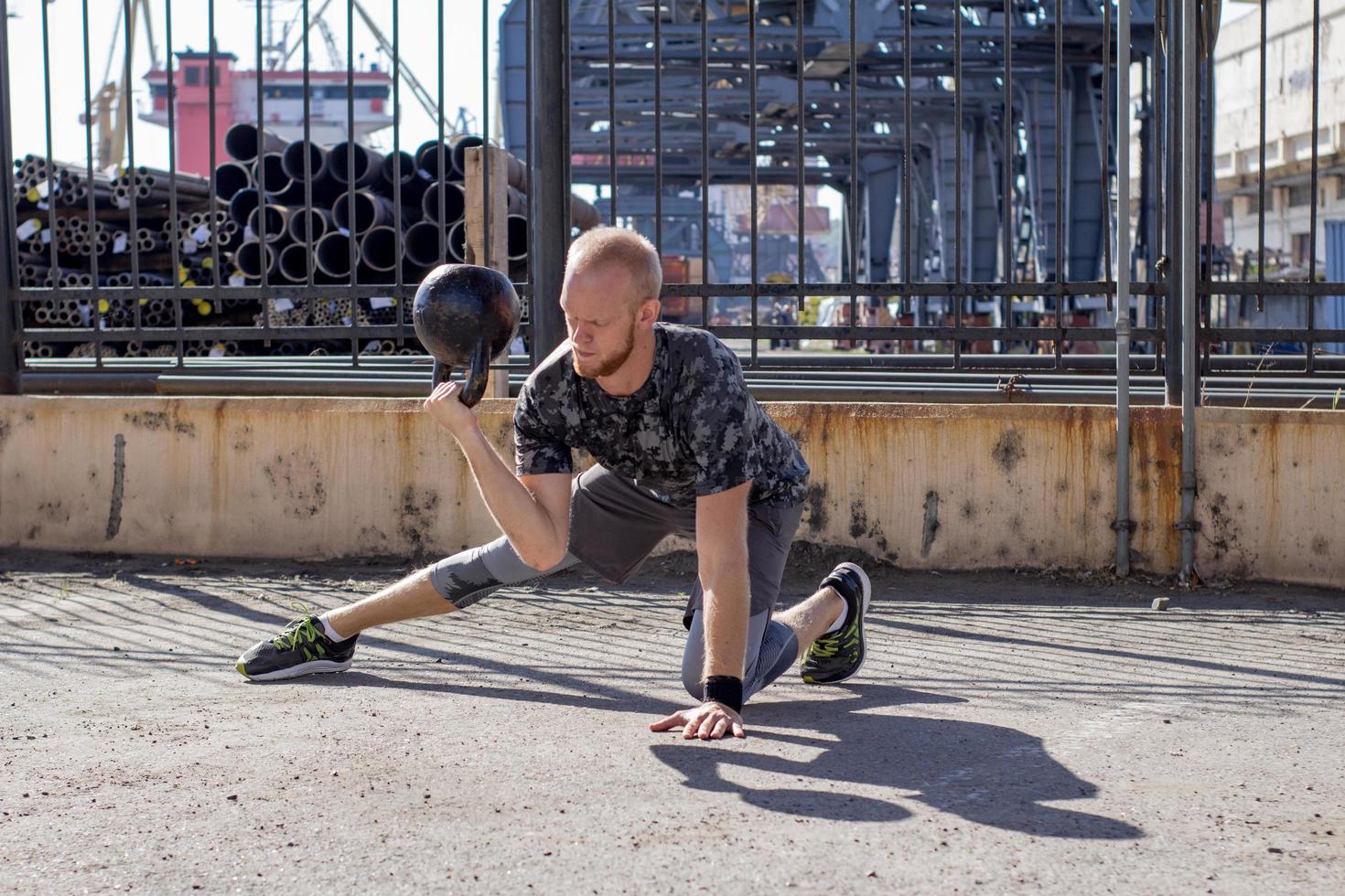 Young bearded male athlete training in industrial zone in sunny day, kettlebells exercises outdoors, urban background photo