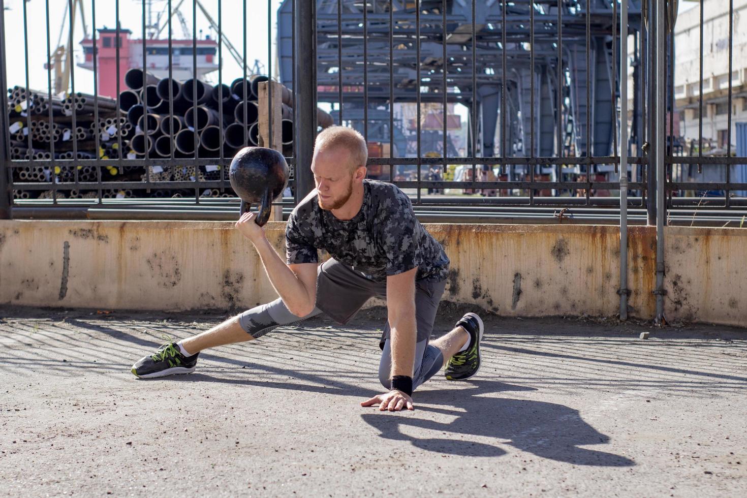 joven atleta masculino barbudo entrenando en zona industrial en un día soleado, ejercicios de campanas al aire libre, fondo urbano foto