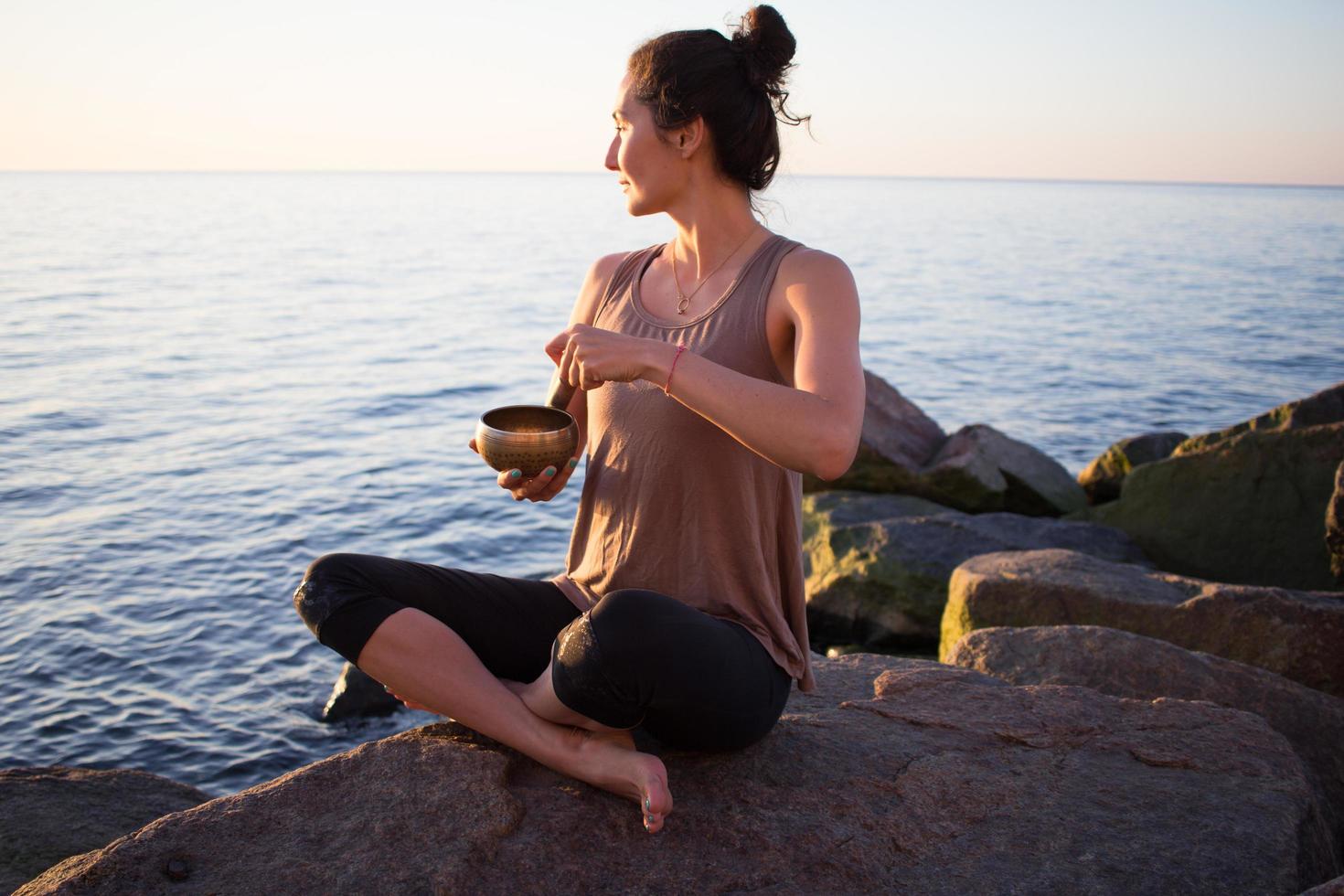 young sporty woman play on Tibetan bowl and sitting on the stones, beautiful asian woman watching sunrise in the sea in yoga pose photo