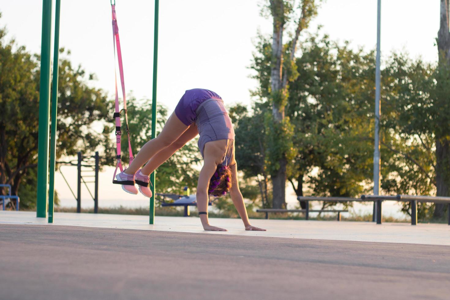 Beautiful fit woman in pink and purple sportwear training on outdoor gym in morning, exercises with suspension straps in park photo