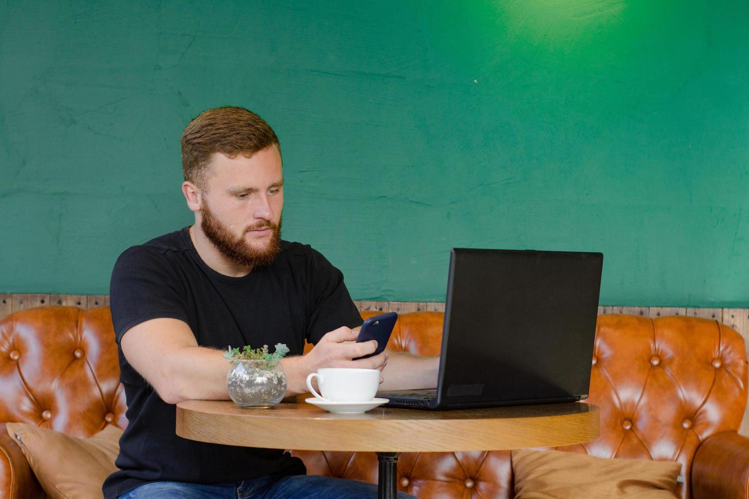 un joven pelirrojo barbudo trabaja en el café con un teléfono celular y una computadora portátil foto