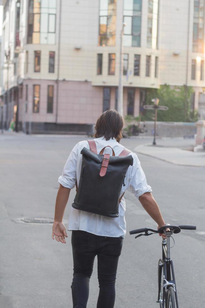 Young bearded man with leather backpack walking with black bicycle photo