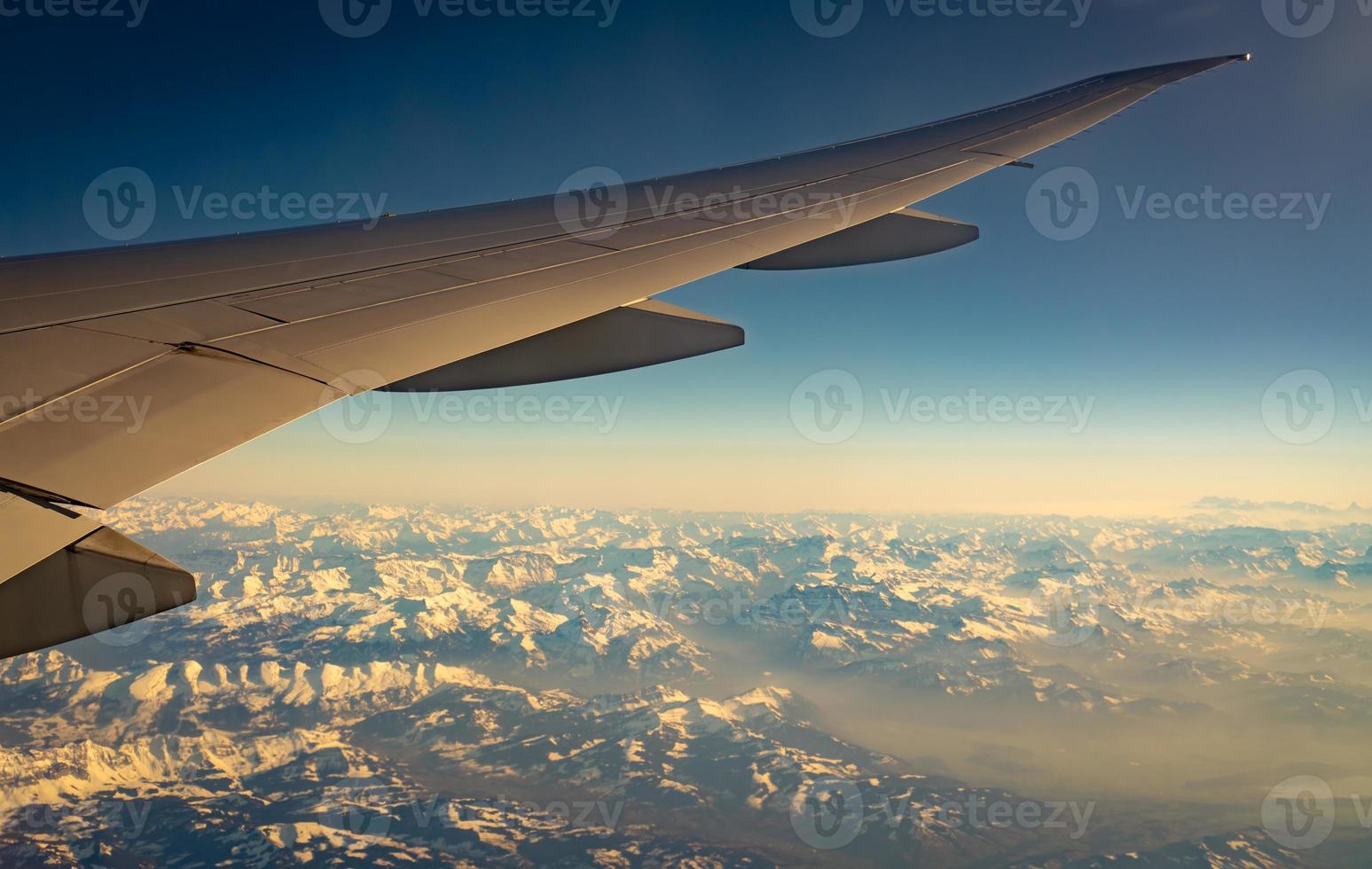 Wing of plane over mountain cover with white snow. Airplane flying on blue sky. Scenic view from airplane window. Commercial airline flight. Plane wing. Flight mechanics concept. International flight. photo